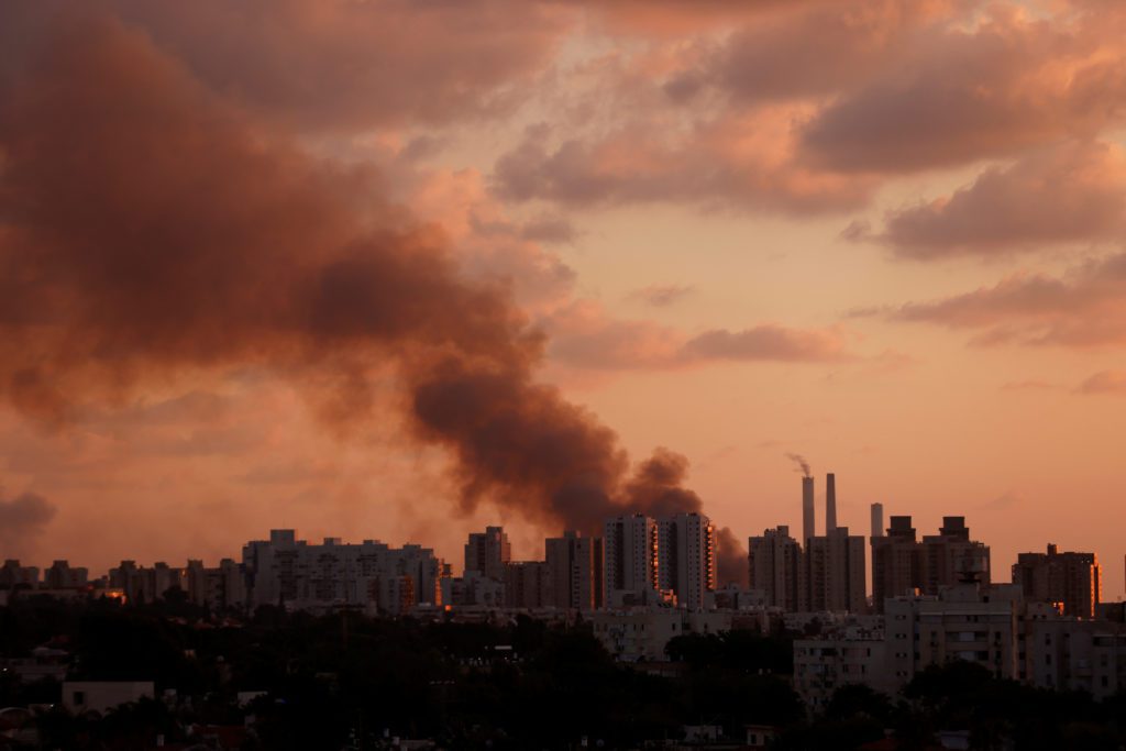 Coluna de fumaça escura sobre cidade ao entardecer, com céu laranja.