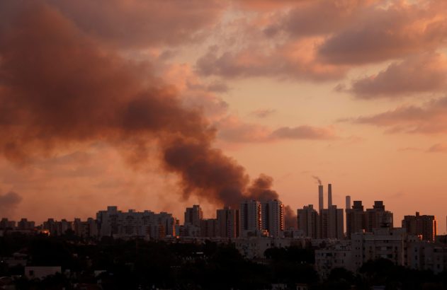 Coluna de fumaça escura sobre cidade ao entardecer, com céu laranja.