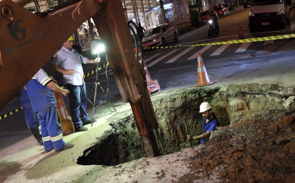 Trabalhadores da construção civil em escavação profunda na rua à noite, com braço de escavadeira visível.
