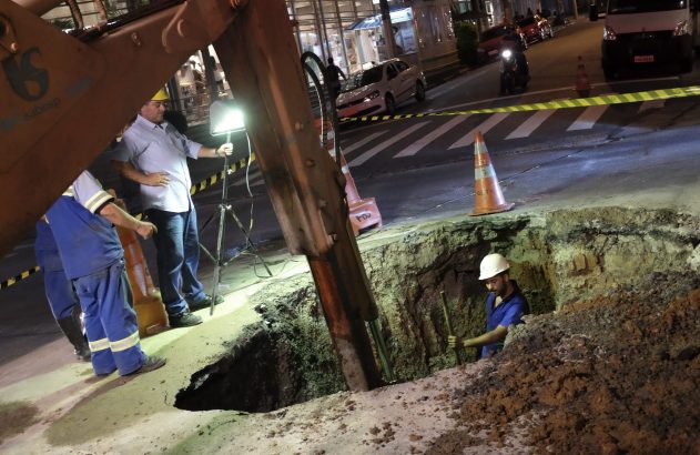Trabalhadores realizam escavação noturna em via pública. Um operário na vala, outro com holofote. Escavadeira e cones.