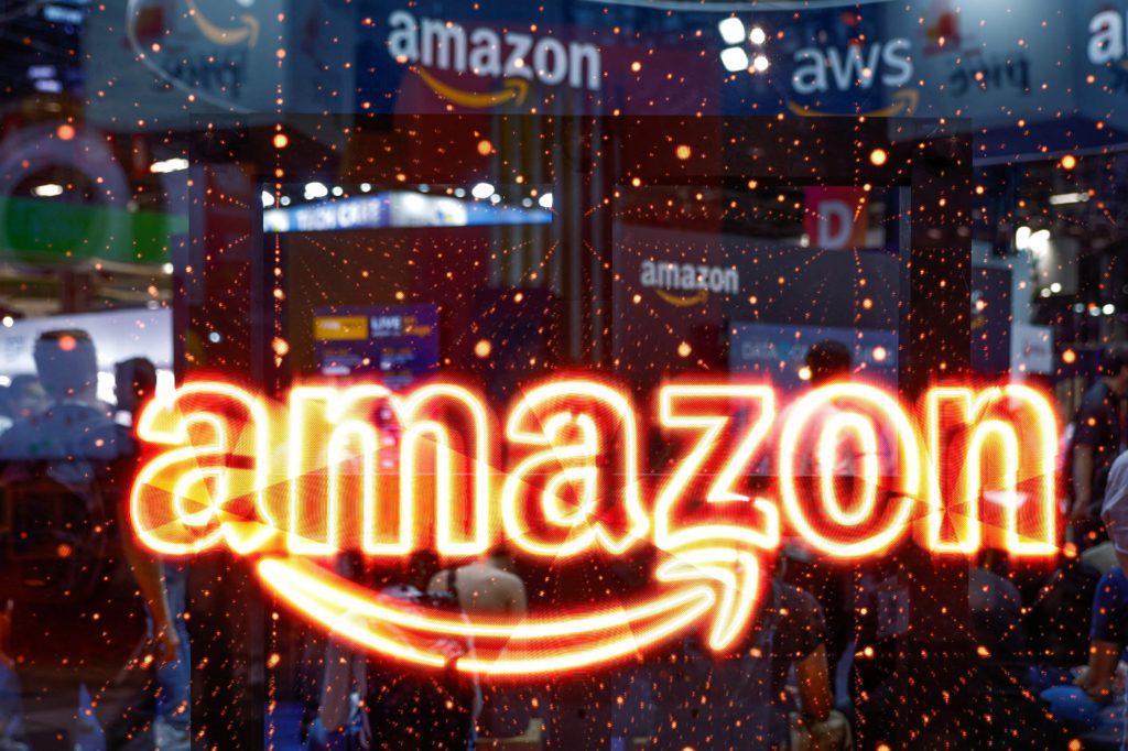 Logo Amazon neon laranja brilhante, com brilhos, em primeiro plano. Fundo de pessoas e estandes em evento Amazon/AWS.