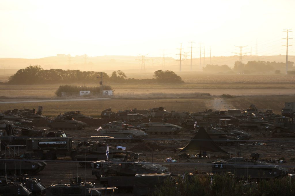 Campo com vários veículos militares avariados. Ao fundo, estrada com veículos em movimento sob céu industrial e nebuloso.