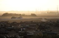 Campo com vários veículos militares avariados. Ao fundo, estrada com veículos em movimento sob céu industrial e nebuloso.