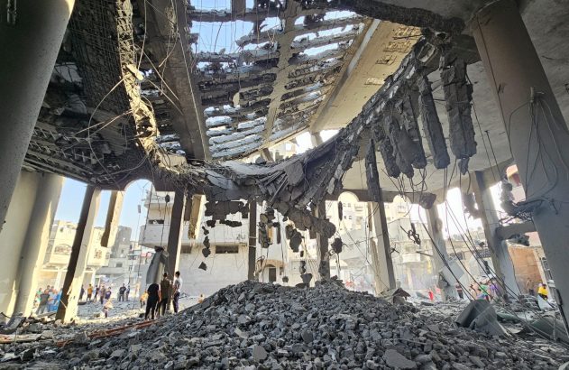Interior de prédio gravemente destruído. Teto e andares colapsados, grande monte de escombros e pessoas observando.