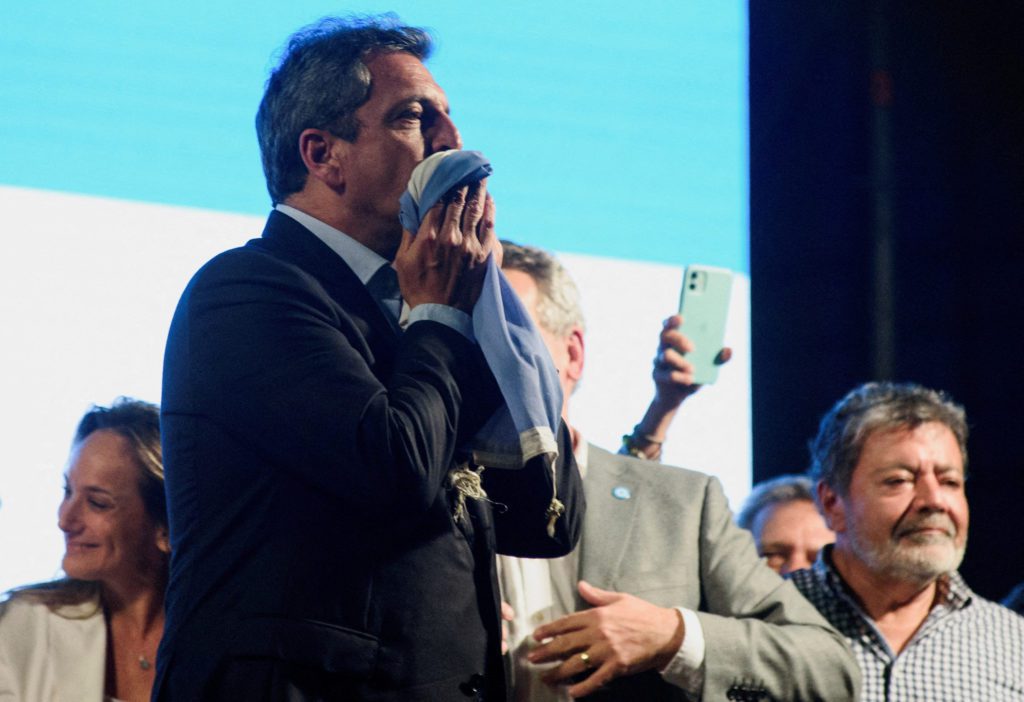 Homem de terno beija um tecido azul e branco no rosto. Pessoas e um celular são visíveis no fundo.