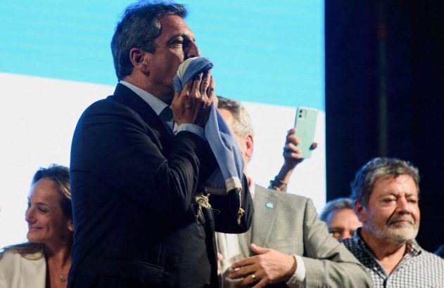 Homem de terno beija um tecido azul e branco no rosto. Pessoas e um celular são visíveis no fundo.