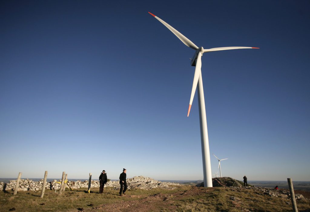 Duas pessoas observam uma turbina eólica em campo aberto sob céu azul.