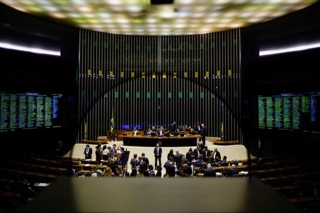 Plenário moderno em sessão política, com pessoas, telas de votação e bandeiras do Brasil.