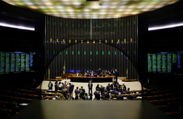 Plenário moderno em sessão política, com pessoas, telas de votação e bandeiras do Brasil.