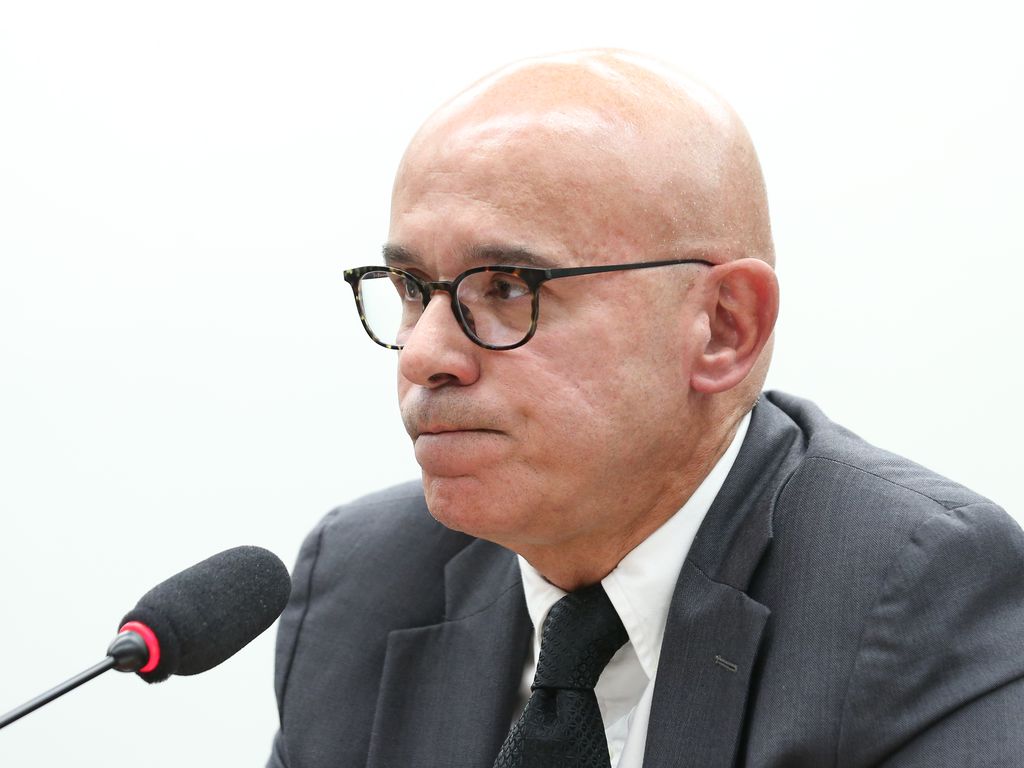 Homem careca de óculos e terno falando ao microfone.