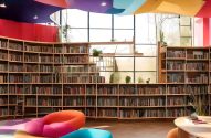 Biblioteca com estantes de livros curvas, janelas amplas, teto colorido e puffs para leitura.