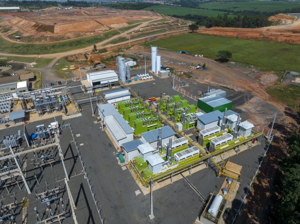 Usina de energia com módulos verdes, subestação elétrica e aterro no horizonte, vista aérea.