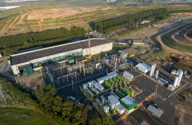 Vista aérea de planta industrial com edifícios, tubulações e tanques em meio a campos e estradas.