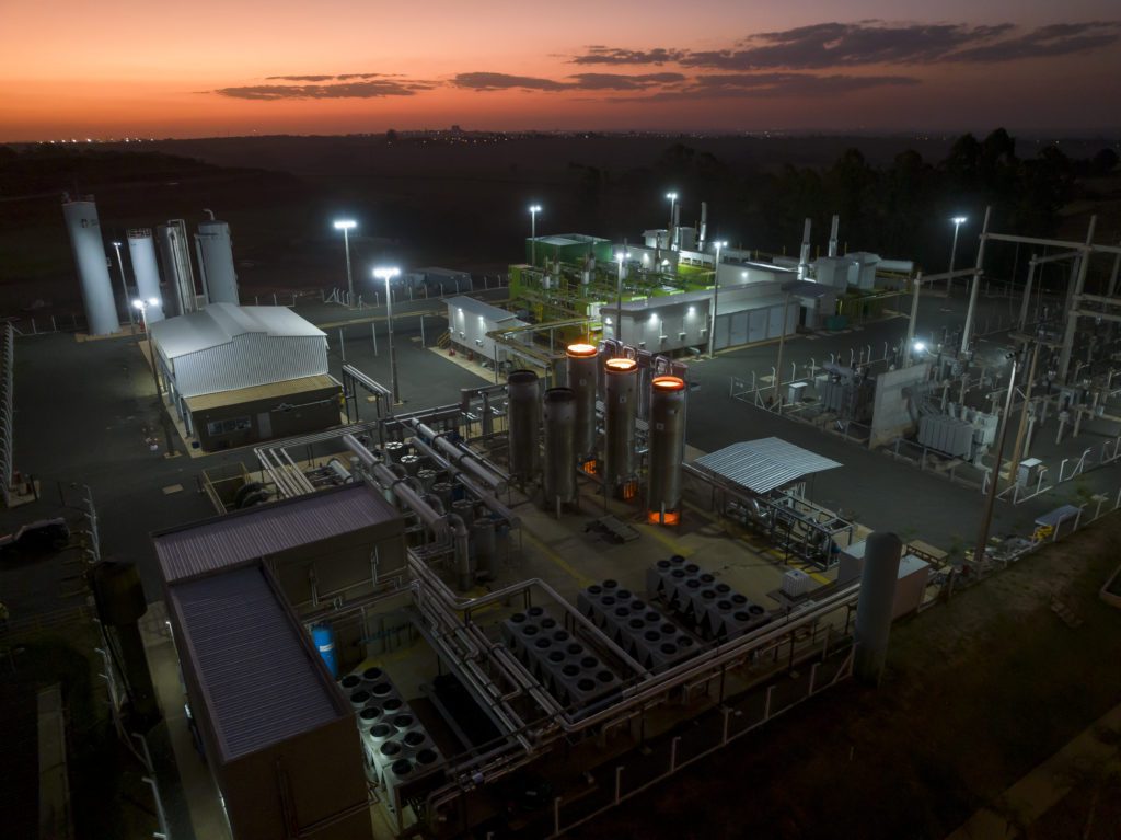 Usina industrial ao anoitecer, com tanques, tubulações, e áreas incandescentes alaranjadas. Céu crepuscular.