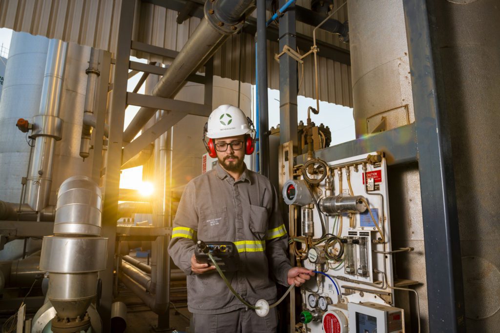 Homem com capacete e fones, operando equipamento em painel industrial. Luz solar ao fundo.