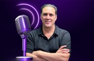 Homem sorrindo, braços cruzados, ao lado de microfone roxo com ondas sonoras. Imagem de podcast ou locução.
