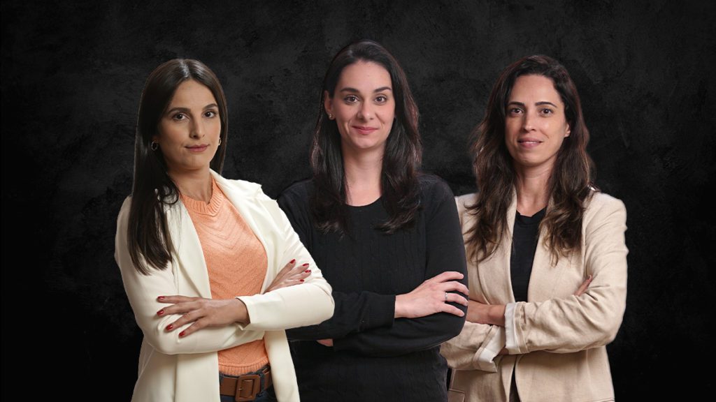 Três mulheres, de pé, sorrindo e com os braços cruzados.