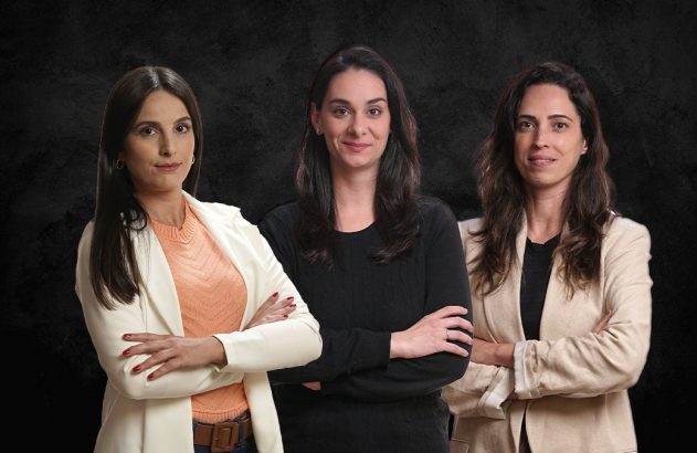 Três mulheres, de pé, sorrindo e com os braços cruzados.