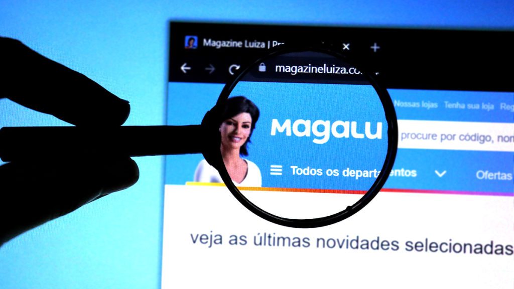 Mão segura lupa sobre site Magalu em monitor, mostrando a assistente virtual Lu.