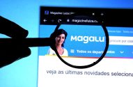 Mão segura lupa sobre site Magalu em monitor, mostrando a assistente virtual Lu.