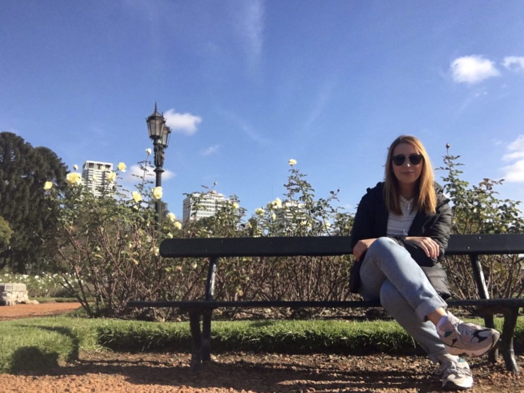 Mulher sentada em banco de parque, óculos escuros, dia ensolarado. Roseiras, poste de luz e prédios ao fundo.