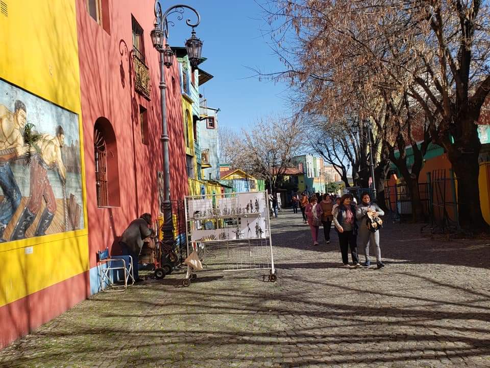 Rua com construções coloridas, mural e pessoas caminhando. Um vendedor expõe produtos em dia ensolarado.