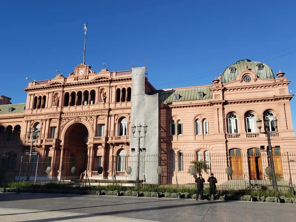Casa Rosada, palácio presidencial rosa da Argentina, em dia claro. Duas figuras caminham em frente.