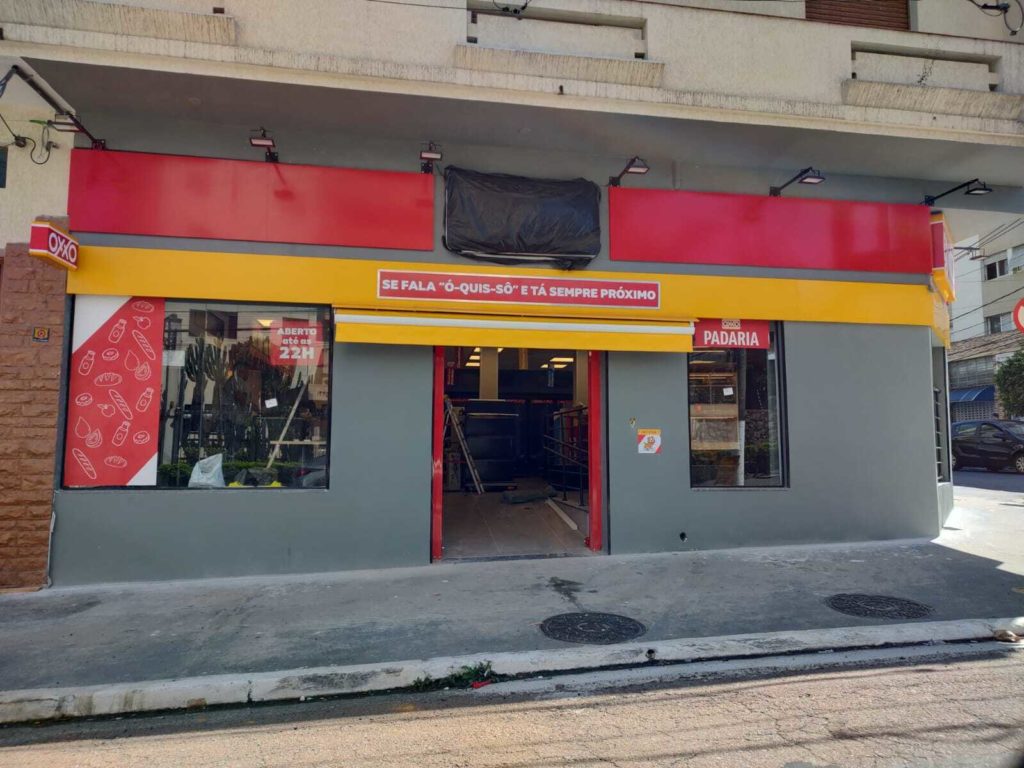Fachada da nova loja OXXO com paredes cinzas e faixas vermelhas e amarelas. Sinais indicam 'ABERTO até as 22H' e 'PADARIA'.