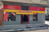 Fachada da nova loja OXXO com paredes cinzas e faixas vermelhas e amarelas. Sinais indicam 'ABERTO até as 22H' e 'PADARIA'.