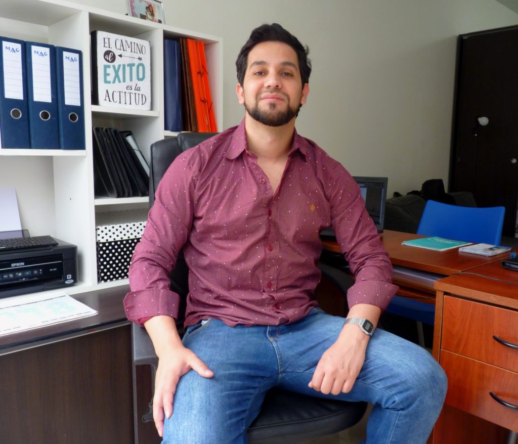 Homem sorridente sentado em escritório com estantes e mesa ao fundo.