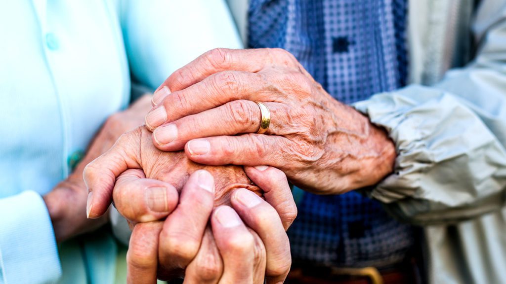 Mãos envelhecidas de duas pessoas se seguram. Uma aliança de ouro em uma das mãos.