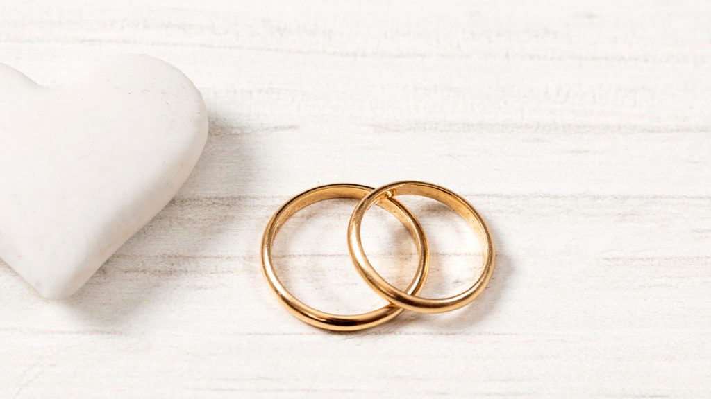 Anéis de casamento dourados entrelaçados sobre madeira, com coração branco à esquerda.