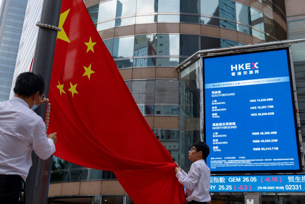 Duas pessoas seguram a bandeira da China em frente a uma tela de cotações financeiras.