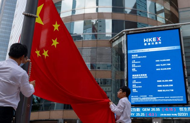 Duas pessoas seguram a bandeira da China em frente a uma tela de cotações financeiras.