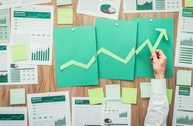 Mão posiciona seta verde de crescimento em painel de gráficos e relatórios financeiros sobre mesa de madeira.