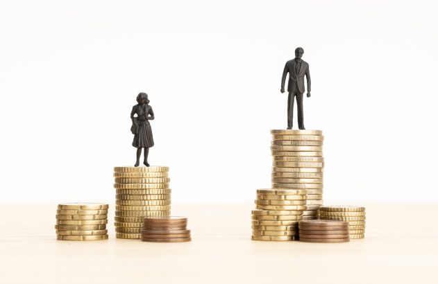 Miniaturas de homem e mulher sobre pilhas de moedas; a do homem é bem maior. Representa a diferença salarial de gênero.