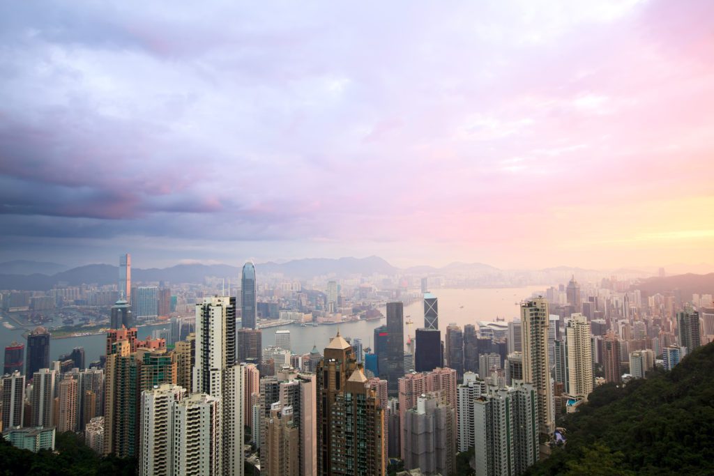 Panorama urbano de Hong Kong ao entardecer com arranha-céus, baía e céu rosa e laranja.