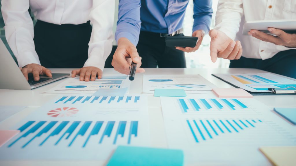 Equipe em reunião analisando gráficos e dados. Um aponta, outro usa notebook, o terceiro tablet.