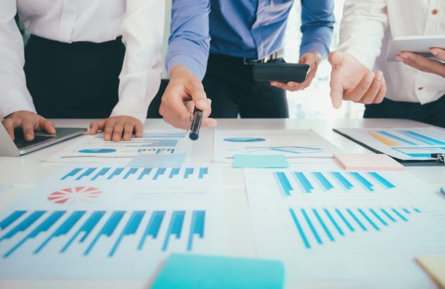 Equipe em reunião analisando gráficos e dados. Um aponta, outro usa notebook, o terceiro tablet.