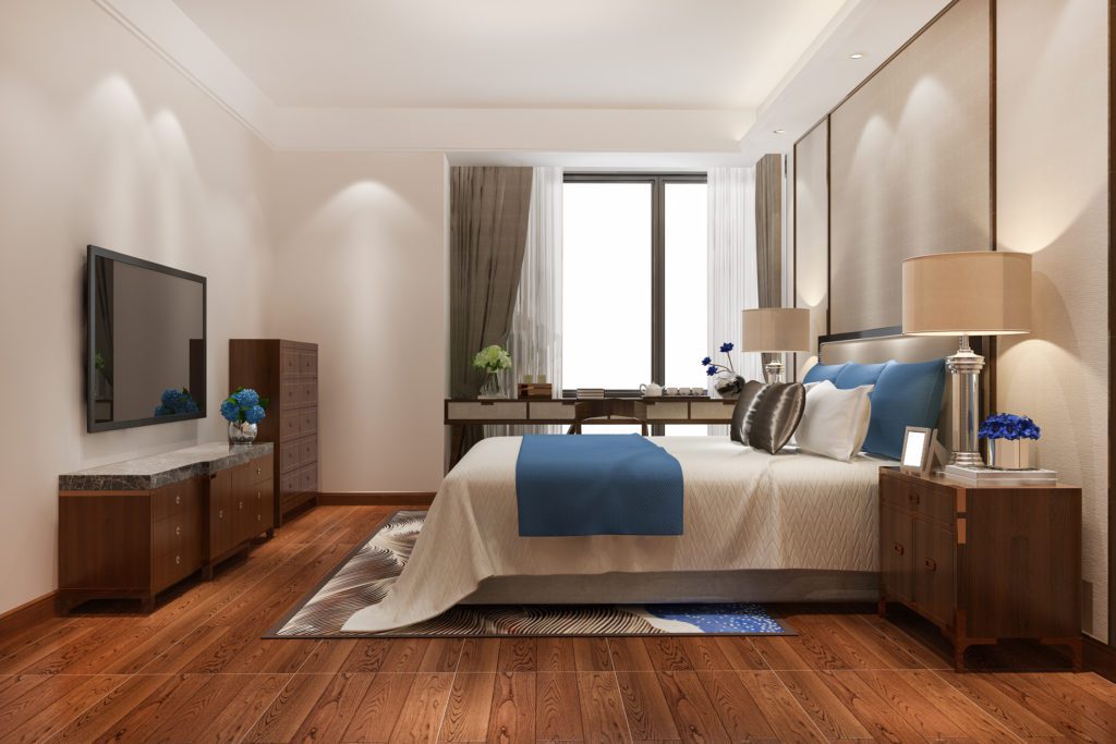 Quarto moderno com cama, travesseiros azuis, TV na parede, móveis de madeira e janela. Bem iluminado, piso de madeira.