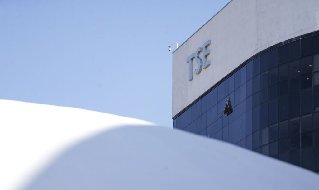 Edifício do TSE com fachada de vidro sob céu azul, e parte de uma estrutura branca curva em primeiro plano.