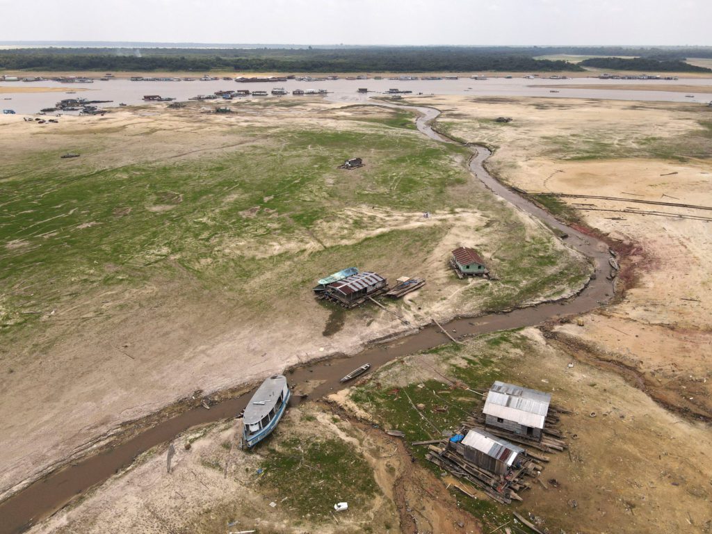 Área de rio em seca extrema com casas e barcos encalhados na margem exposta. Um pequeno riacho serpenteia pelo leito seco.
