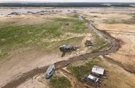 Área de rio em seca extrema com casas e barcos encalhados na margem exposta. Um pequeno riacho serpenteia pelo leito seco.