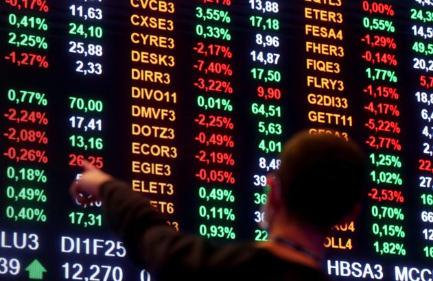 Pessoa apontando para tela digital de bolsa de valores com cotações e percentuais de ações.