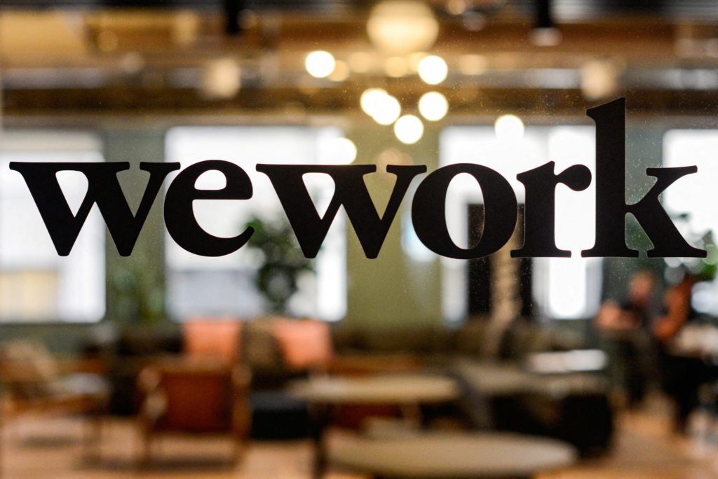 A palavra 'wework' em preto sobre um vidro. Fundo desfocado de um escritório.