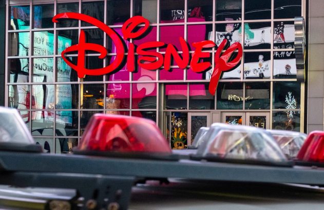 Fachada da Disney com logo vermelho e luzes de emergência de viatura policial em primeiro plano.