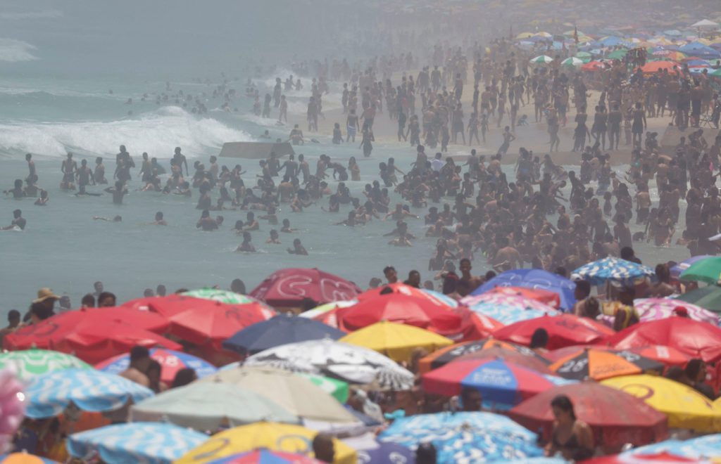 Praia lotada com guarda-sóis coloridos e muitas pessoas no mar e na areia.