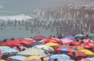 Praia lotada com guarda-sóis coloridos e muitas pessoas no mar e na areia.