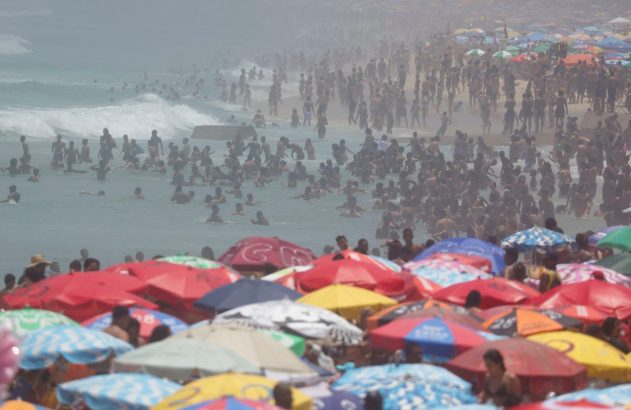 Praia lotada com guarda-sóis coloridos e muitas pessoas no mar e na areia.