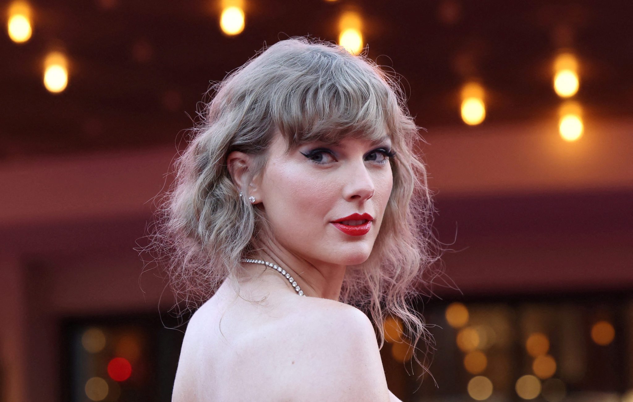 Taylor Swift olhando para trás, cabelo loiro ondulado curto e batom vermelho.